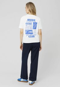T-shirt en coton blanc avec un design graphique bleu présentant des illustrations de nourriture et de boissons. Associé à un pantalon marine et des baskets aux accents bleus.