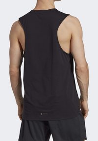 Homme portant un débardeur sportif noir sans manches et un short noir, vu de dos sur un fond clair uni.