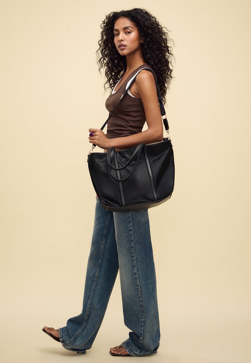 Schwarze Lederhandtasche mit zwei Tragegriffen oben, schlankem Design und matter Oberfläche. Kombiniert mit weit geschnittenen blauen Jeans und einem braunen Tanktop.