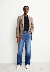 Bruine wollen blazer over een zwart polo-shirt, gecombineerd met wijde blauwe jeans en crèmekleurige sneakers. Geweven stof en een getailleerde pasvorm zijn zichtbaar.