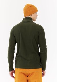 Maglia verde a maniche lunghe con colletto alto, realizzata in materiale morbido. Abbinata a un berretto lavorato a maglia arancione e pantaloni arancioni. Vista posteriore mostrata.