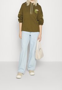Persona vestida con sudadera verde oliva con un gráfico, jeans de pierna ancha de color azul claro, zapatillas beiges y sosteniendo un pequeño bolso de mano beige y esponjoso.