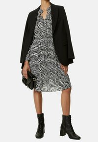 Vestido negro con flores, con cuello y detalle de lazo, superpuesto a un blazer negro. Combinado con botines negros con cordones y un bolso negro.