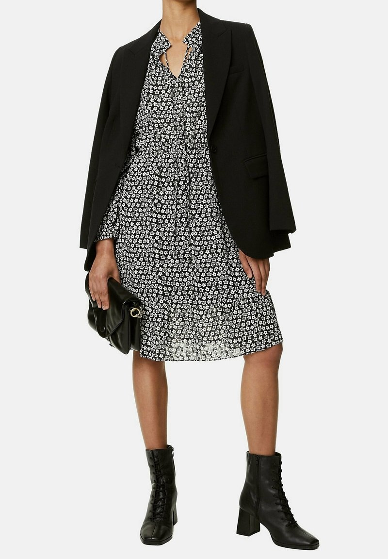 Vestido negro con flores, con cuello y detalle de lazo, superpuesto a un blazer negro. Combinado con botines negros con cordones y un bolso negro.
