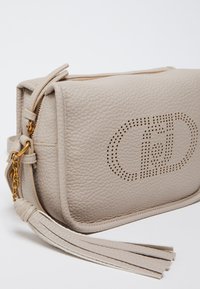 Sac bandoulière en cuir crème avec logo perforé, quincaillerie dorée et tirette de fermeture à glissière frangée. Forme rectangulaire avec surface texturée.