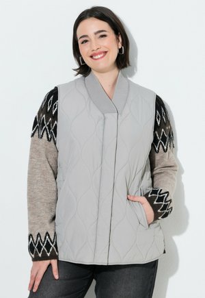 Donna sorridente che indossa un gilet trapuntato grigio chiaro sopra un maglione nero e beige a fantasia, con una mano nella tasca del gilet e pantaloni scuri.