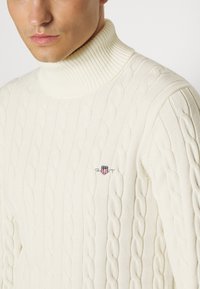 Cable-knit trui in gebroken wit, met een geribde hoge kraag. Inclusief klein merkembleem aan de linkerkant. Zacht, gestructureerd materiaal.