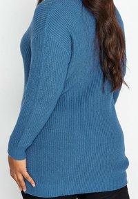 Blå stickad tröja med långa ärmar, med ribbat mönster och en avslappnad passform. Sömmen är något längre bak.