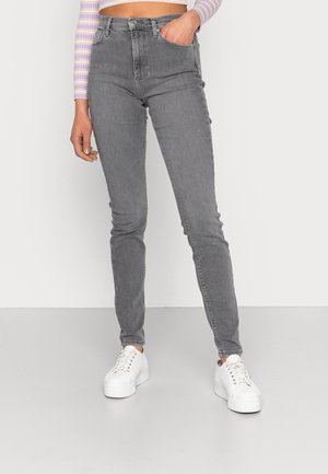 Femme portant un jean skinny gris taille haute, des baskets blanches à plateforme et un top rayé lavande à manches longues raccourci, une main dans la poche.
