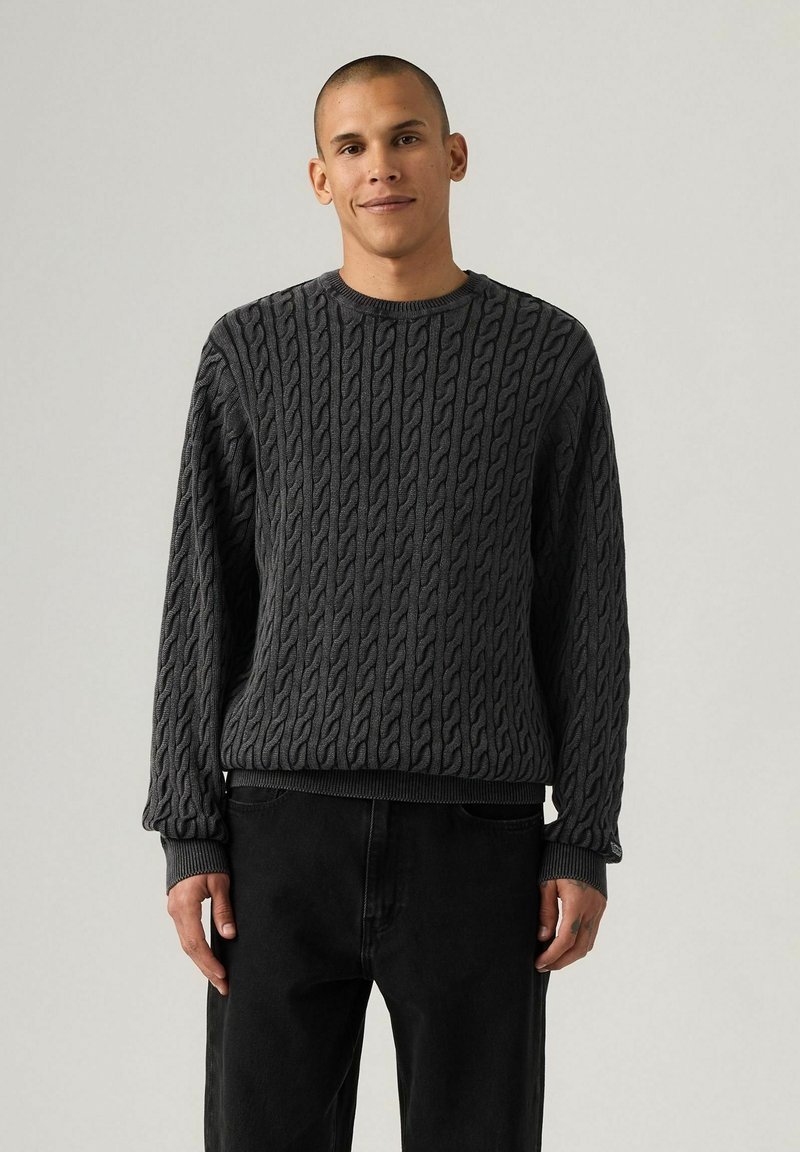 Dark gray knitted sweater featuring a cable pattern, crew neckline, and ribbed cuffs and hem. Paired with black trousers.