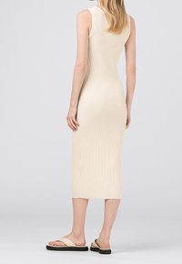 Stickad, ribbad midiklänning i ljusbeige, ärmlös design med en figurnära silhuett. Kombinerad med neutrala slidesandaler, som visar upp en minimal estetisk.