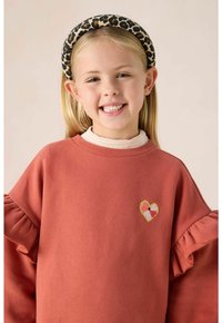 Rostfarbener Pullover mit rüschengeschnittenen Ärmeln, der auf der Brust eine herzförmige Blumenstickerei zeigt. Wird mit einem Stirnband im Leopardenmuster getragen.