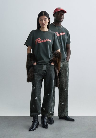 Två personer i mörkgröna t-shirts med texten "Passion", kombinerade med broderade jeans och svarta ankelstövlar. Den ena bär en pälsdetalj.