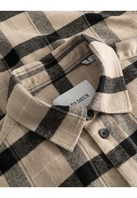 Beige flanellskjorte med et svart og kremmønstret rutemønster. Har krage med knapper og mørke knapper, og viser en myk tekstur.