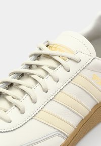 Primo piano di una sneaker Adidas bianca con strisce beige, testo "Spezial" e suola in gomma beige che mostra dettagli dei lacci e delle cuciture.