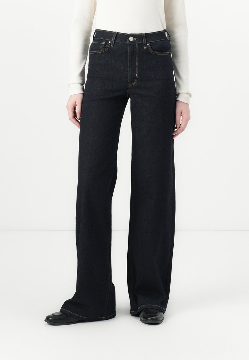 Weite, dunkle Jeans mit hoher Taille, ausgestattet mit klassischer Naht und fünf Taschen, kombiniert mit einem eng anliegenden, weißen Langarmoberteil.