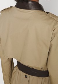 Beżowy płaszcz trenczowy z brązowym skórzanym kołnierzem, talią w pasie i guzikami ozdobnymi. Posiada warstwowy design pleców z gładką fakturą.