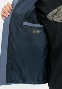 Vandafvisende og pletbestandig jakke med en marineblå indvendig side og grå accenter, der har en inderlomme og plejeikoner.