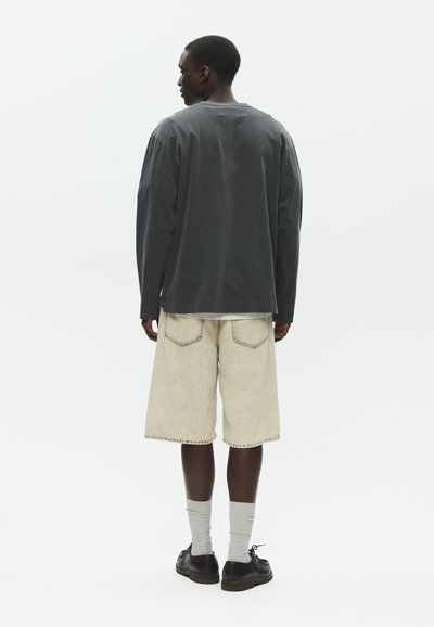 Homme debout, tourné de dos, portant une chemise à manches longues foncée, un short beige arrivant aux genoux, des chaussettes grises et des chaussures noires sur un fond uni.