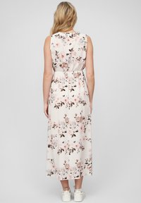 Florale maxi jurk in lichtcrème met roze en bruine bloemenpatronen. Mouwloos ontwerp met een elastische tailleband. Witte sneakers zichtbaar.