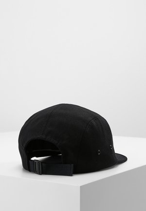 Czarna regulowana czapka z paskiem spoczywająca na białym sześcianie na jednolitym jasnoszarym tle.