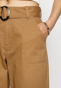 Bruna högmidjade byxor med vida ben, som har en stor cirkulär bältesknäppning, sidoficka och synliga sömdetaljer.