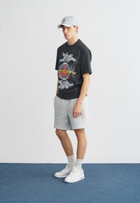 Μαύρο γυναικείο t-shirt με πολύχρωμο σχέδιο, γκρι αθλητικές βερμούδες, λευκά αθλητικά παπούτσια και ανοιχτό γκρι καπέλο μπροστά σε ουδέτερο φόντο.