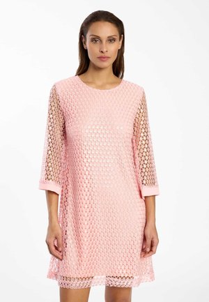 Femme portant une robe rose en dentelle longueur genou avec des manches trois-quarts transparentes, debout devant un fond blanc uni.