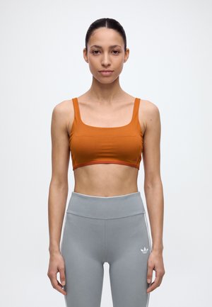 Une femme se tient face à l’objectif, vêtue d’une brassière de sport orange et d’un legging gris, devant un fond clair uni.