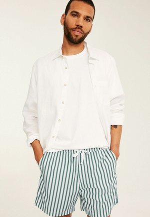 Mand med skæg iført en hvid skjorte, hvid t-shirt og grøn-hvide lodrette stribede shorts med hænderne i lommerne.