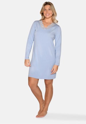 Femme souriante portant une chemise de nuit bleu clair à manches longues avec un col en V bordé de dentelle, debout pieds nus devant un fond uni.