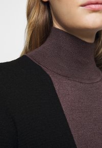 Kvinna i lila ribbad polotröja under en svart strukturerad cardigan, med långt ljusbrunt hår synligt.