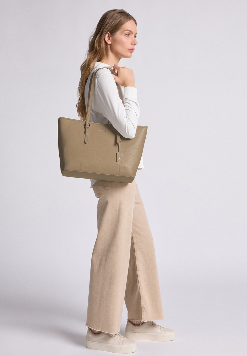 Femme portant un pantalon large beige et un haut blanc, portant un grand sac cabas en cuir taupe sur son épaule, se tenant contre un fond uni.