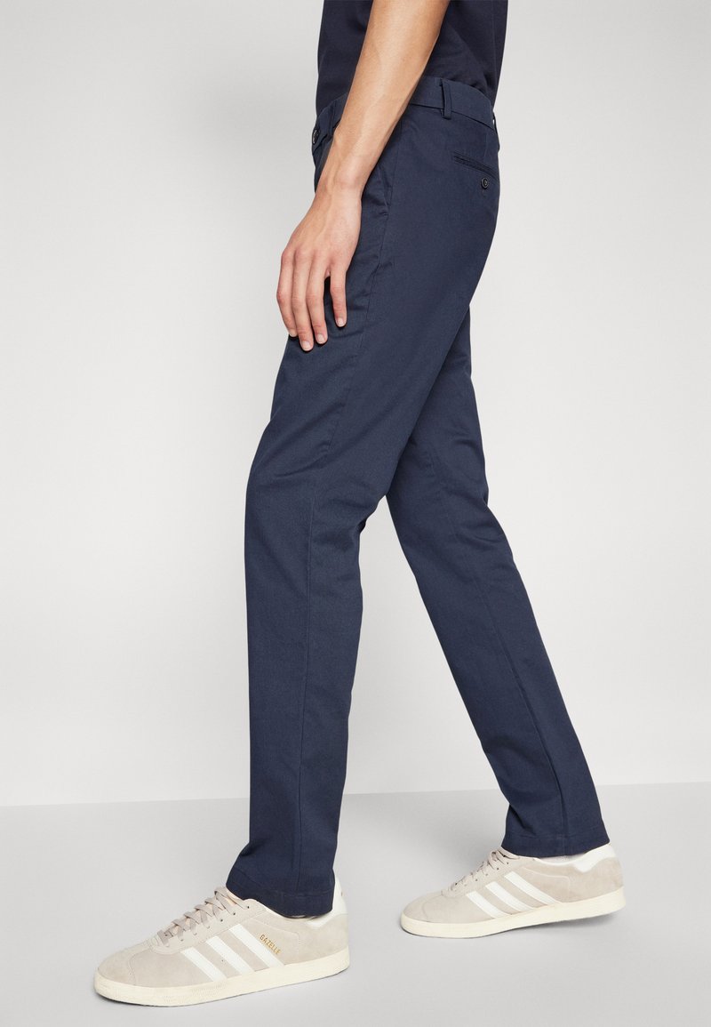Pantalon bleu marine en coton, avec une coupe slim et un devant plat, associé à des baskets beiges avec des rayures blanches.
