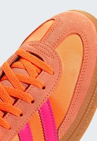 Orange athletic shoe with suede and satin sections, featuring pink serrated stripes, textured laces, and a gum rubber sole.