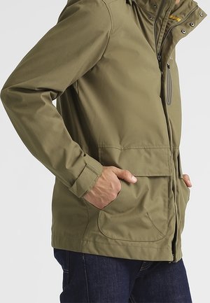 Osoba w oliwkowo-zielonej kurtce z zamkiem błyskawicznym i zatrzaskami, z rękami w przednich kieszeniach, w połączeniu z ciemnoniebieskimi dżinsami.