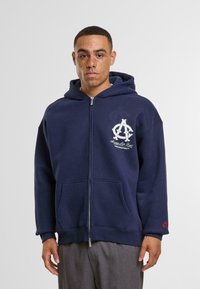 Námornícky modrá mikina na zips s predným vreckom, bielym logom na hrudi a červeným akcentom na rukáve; vyrobená z jemného, textúrovaného materiálu.