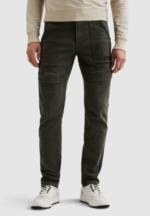 Hombre con pantalones cargo de color verde oliva oscuro con múltiples bolsillos, suéter beige y zapatillas blancas, de pie sobre un fondo liso.