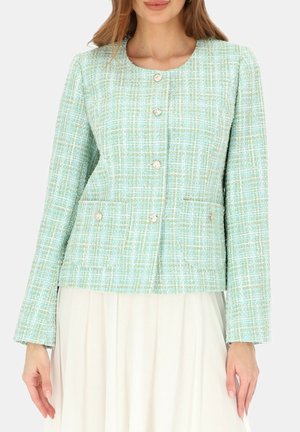 Kobieta ubrana w jasnoniebiesko-zieloną marynarkę z tweedu z perłowymi guzikami, na białej plisowanej spódnicy.
