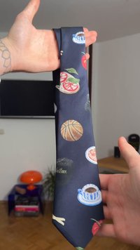 Σκούρο μπλε γραβάτα με διάφορες πολύχρωμες εικόνες φαγητού και ποτών, όπως φράουλες, μπάλες μπάσκετ και κούπες καφέ, κατασκευασμένη από λείο ύφασμα.