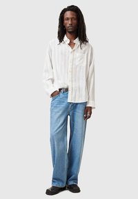 Mężczyzna z długimi dredami, ubrany w białą koszulę z długim rękawem, jasnoniebieskie, szerokie dżinsy, czarne buty oraz srebrny naszyjnik na jednolitym tle.