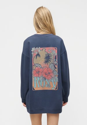 Plavolaska ženska, obrnjena stran, nosi preveliko mornarsko modro majico z napisom "ROXY" na hrbtu ter tropskim sončnim zahodom, kokosovimi palmami in cvetjem.