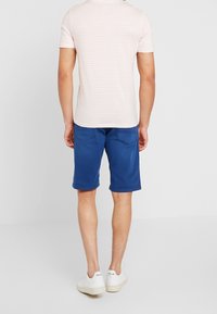 Miesten raidallinen vaaleanpunainen t-paita yhdistettynä sinisiin shortsialusiin. Valkoiset lenkkarit näkyvät. Puuvillamateriaali, rento istuvuus, hienovaraiset ompeludetailit.