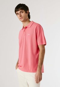 Jeune homme portant un polo rose à manches courtes et un pantalon blanc, debout avec une main dans la poche sur un fond blanc uni.