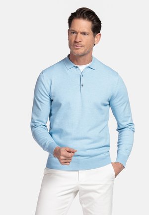 Man met kort bruin haar, gekleed in een lichtblauw poloshirt met lange mouwen over een wit ondershirt en witte broek, staand met één hand in de zak.