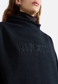 Pullover in pile blu navy oversize con colletto alto, presenta dettagli del logo in rilievo al centro. Texture morbida, senza chiusure visibili.