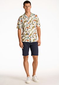 Kortærmet skjorte med knaplukning, fremstillet i et lyst stof med ananas- og bladmønstre, parret med marineblå shorts og hvide sneakers.