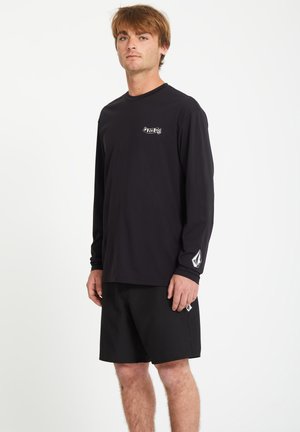 Jeune homme portant un t-shirt noir à manches longues et un short assorti avec des logos blancs, debout devant un fond blanc uni.