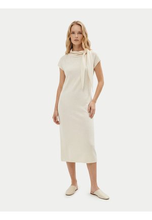 Frau stehend, trägt ein cremefarbenes Midikleid mit kurzen Ärmeln, einem gebundenen Schal am Hals und passenden Slip-On Schuhen.