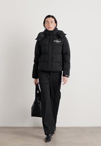 Zwarte pufferjack met ritssluiting en capuchon, voorzien van een logo op de borst, gedragen met zwarte broek en een zwarte tote tas.
